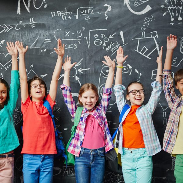 Group of happy schoolchildren standing in front of the blackboard with hands up